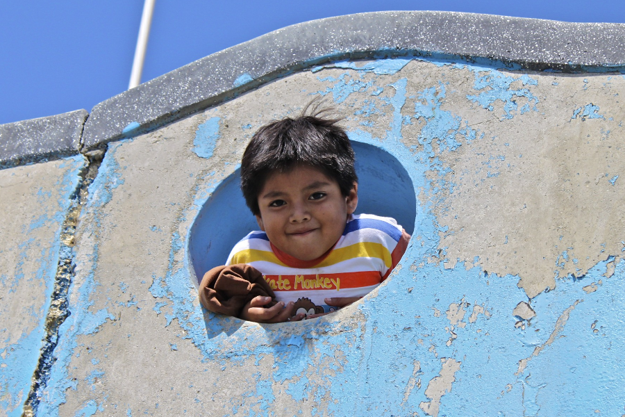 Niño sonriendo, saliendo de una abertura de forma redonda en un muro de concreto, con un cielo azul atrás