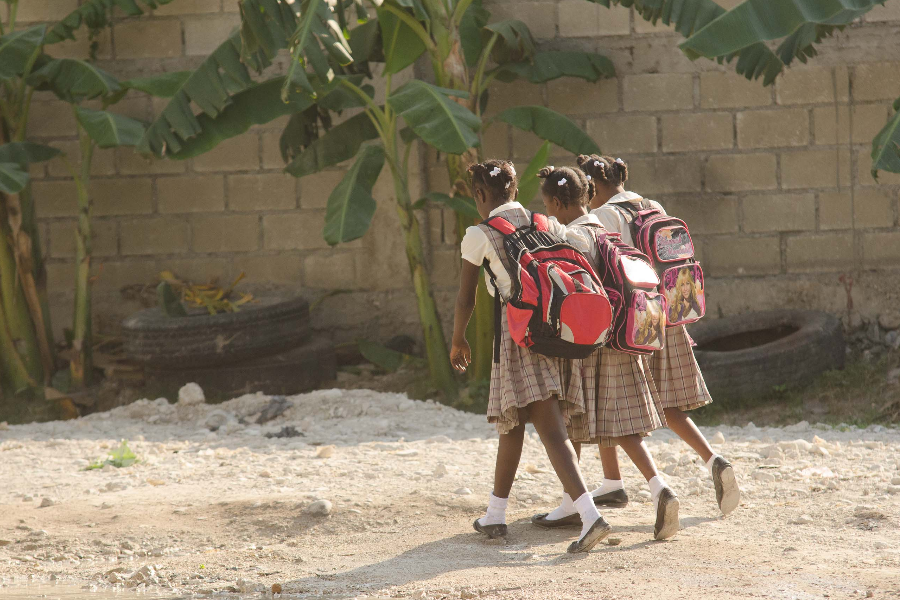 Tres chicas jóvenes que caminan en uniformes escolares haitianos, con mochilas, en un camino sin pavimentar, con algunos árboles de plátano en el fondo