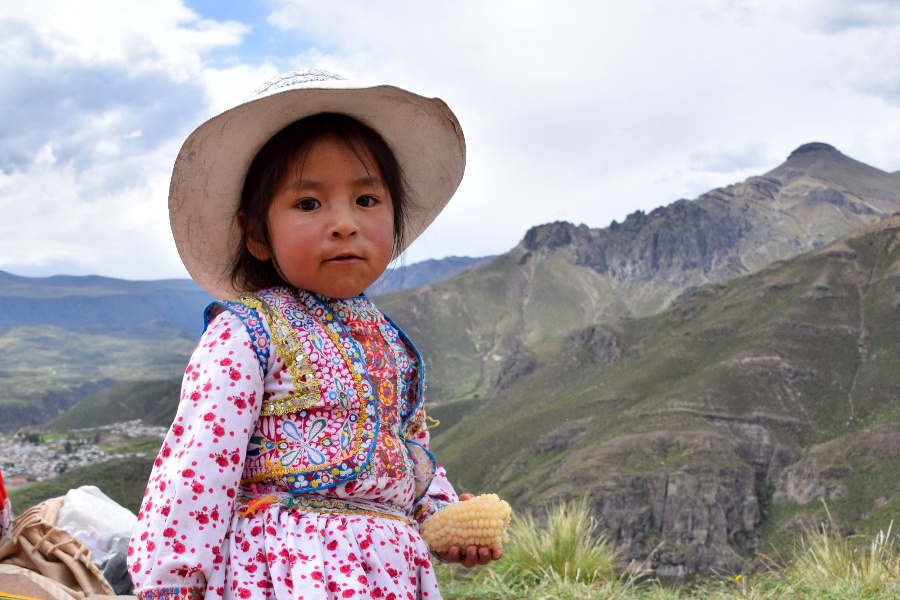 Joven peruana vestida con una camisa rosa de manga larga y un sombrero, sosteniendo un maíz pequeño. En la parte posterior de la imagen, hay montañas y un cielo azul.